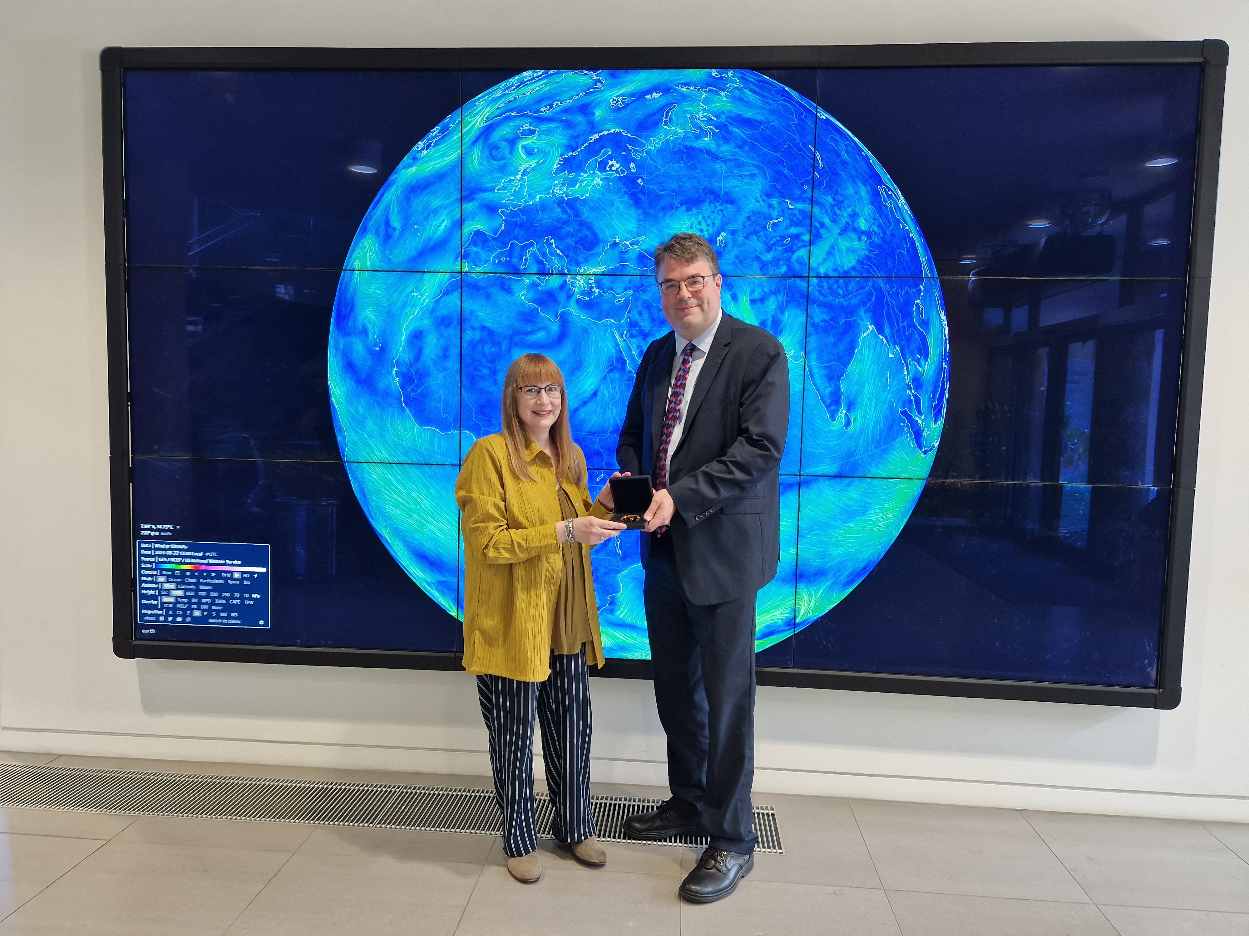 Sharon Ann receiving her medal from Tom Grinyer at IOP London with a live weather map of winds around the world in the background.