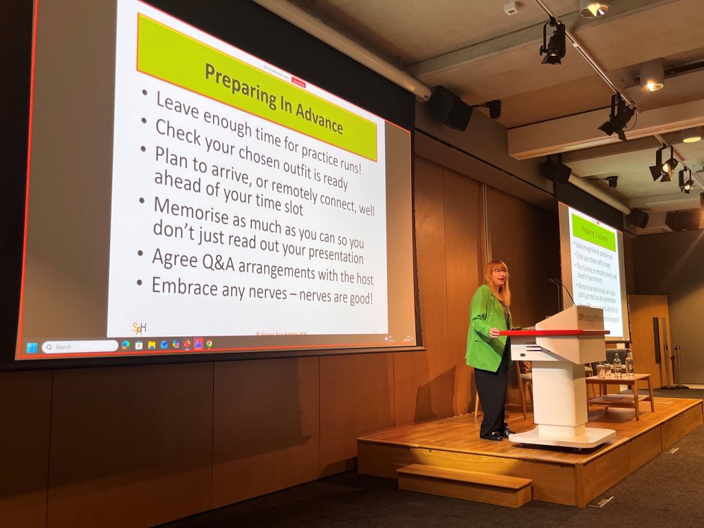 Sharon Ann on a stage behind a podium wearing a green jacket giving a talk in a large lecture room at the Institute of Physics HQ in London.